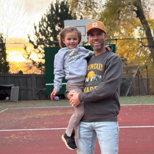 Man holding a child on an outdoor basketball court with trees and sunset in the background