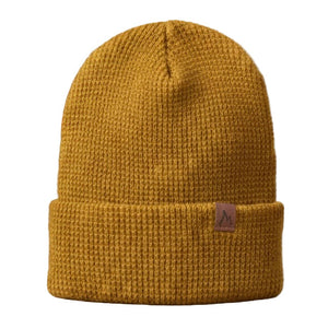 Yellow knit beanie with a brand logo on a wooden surface with a fireplace in the background