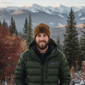 Man wearing a green puffer jacket and brown beanie standing in a snowy landscape with mountains and trees.